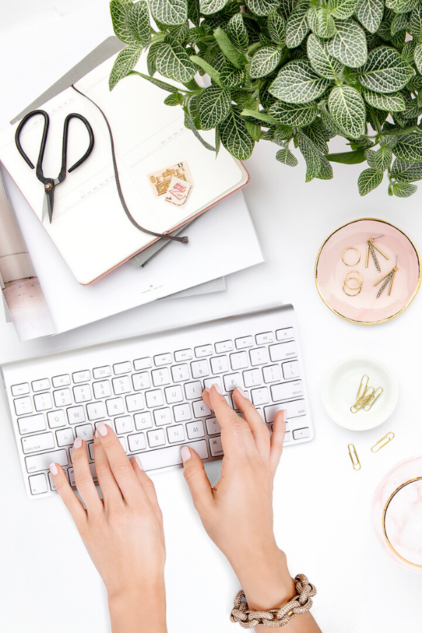 A light and airy color scheme work desk with plant, planner, paper clips and a womans well manicured hands typing on a keyboard.